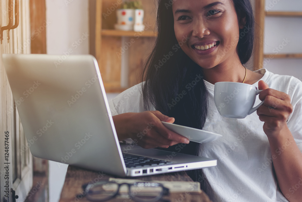 Beautiful hipster woman smiling sitting near window while looking at open laptop computer on table and holding white mug at cafe. Beautiful hipster woman smiling sitting near window while looking at open laptop computer on table and holding white mug at cafe.