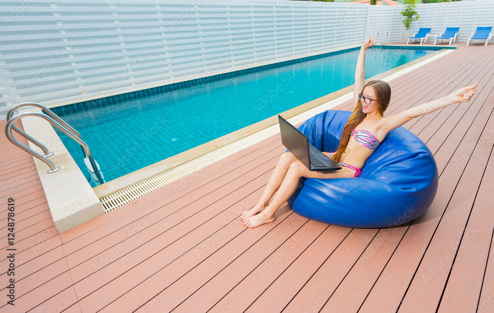 Freelance businesswoman. Young smiling woman in bikini and glasses with laptop  and rised hands , sitting on blue beanbag. Swimming pool on background.