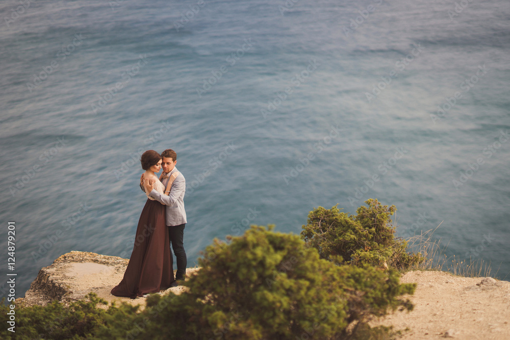 Romantic and stylish caucasian couple standing on the background of spectacular sea view. Love, relationships, romance, happiness concept.