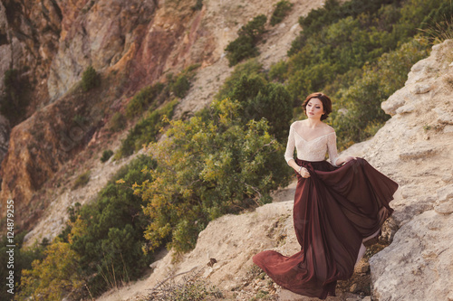 Portrait of a beautiful young caucasian stylish woman in evening gown standing on the background of spectacular nature  rock and trees  in sunlight
