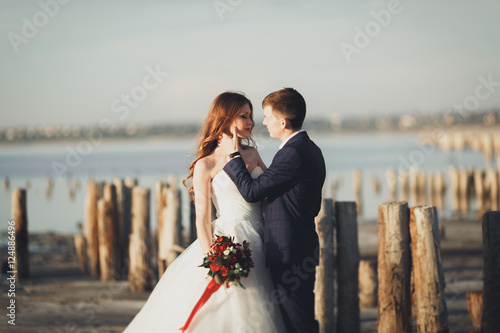 Wedding couple, groom, bride with bouquet posing near sea on sunset