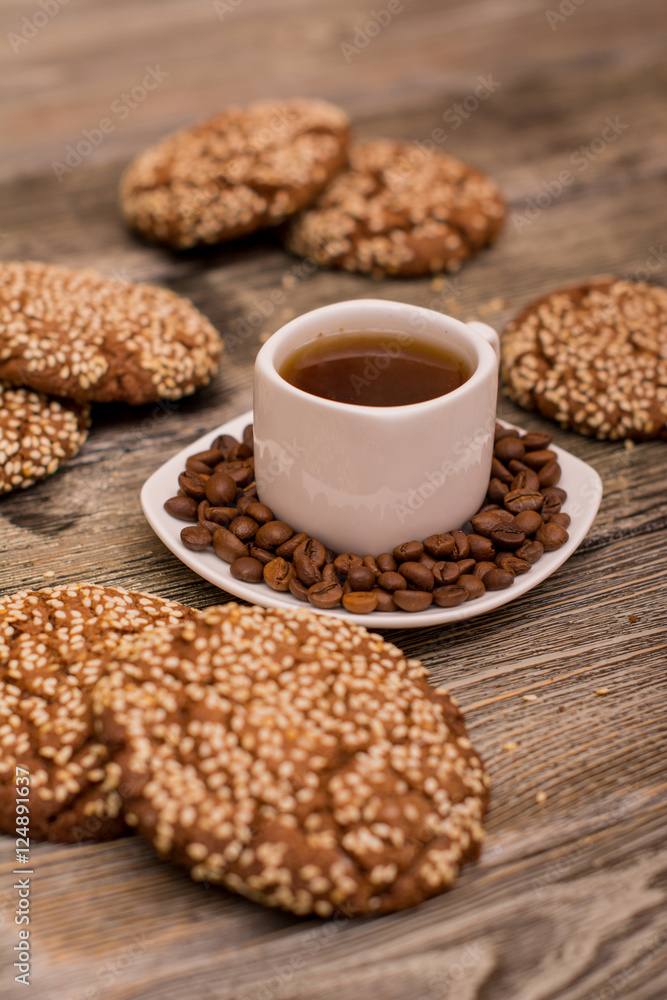 Small white cup of coffee, roasted coffee beans, cookies with sesame seeds on wooden background Small white cup of coffee, roasted coffee beans, cookies with sesame seeds on wooden background