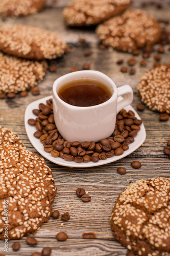 Small white cup of coffee, roasted coffee beans, cookies with sesame seeds on wooden background