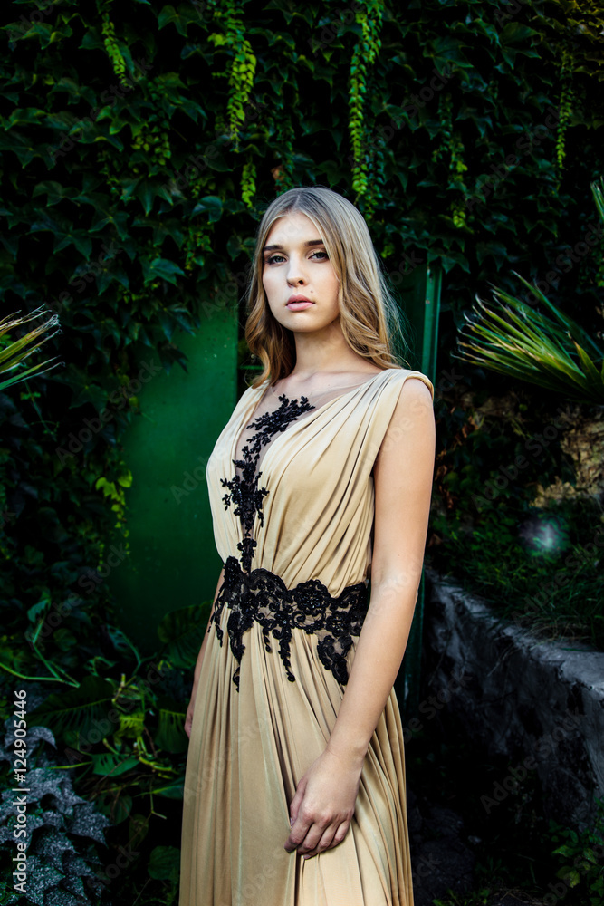 Fototapeta premium Beautiful girl in silk dress posing by wall covered with ivy