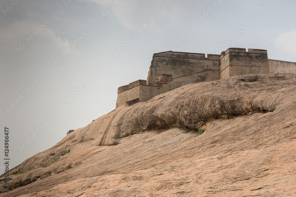 Dindigul, India - October 23, 2013: Brown-beige boulder with the ramparts of the historic Dindigul Rock fort under light blue sky. Brown-beige as dominant color.