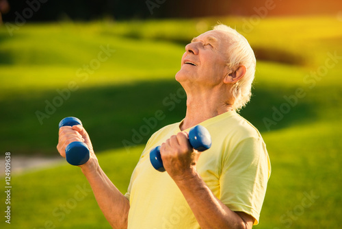 Senior man holding dumbbells. Person smiling and looking up. Strength and concentration. Motivated by the goal.