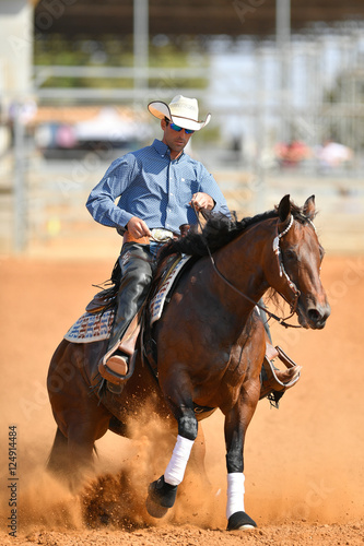 The side view of a rider in cowboy chaps, boots and hat on a horseback running ahead and stopping the horse in the dust. © PROMA