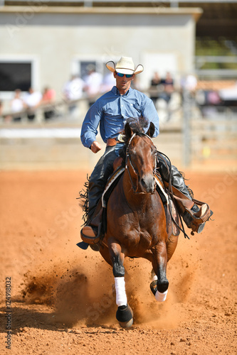 The front view of a rider in cowboy chaps, boots and hat on a horseback running ahead and stopping the horse in the dust.