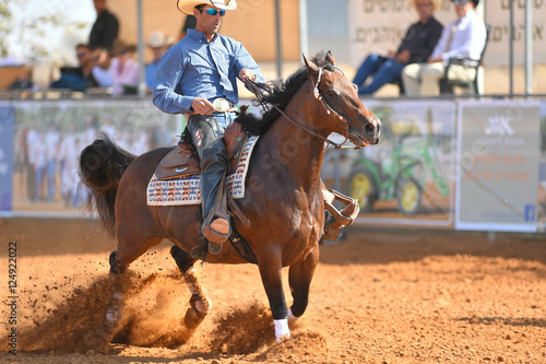 The side view of a rider in cowboy chaps, boots and hat on a horseback running ahead and stopping the horse in the dust.