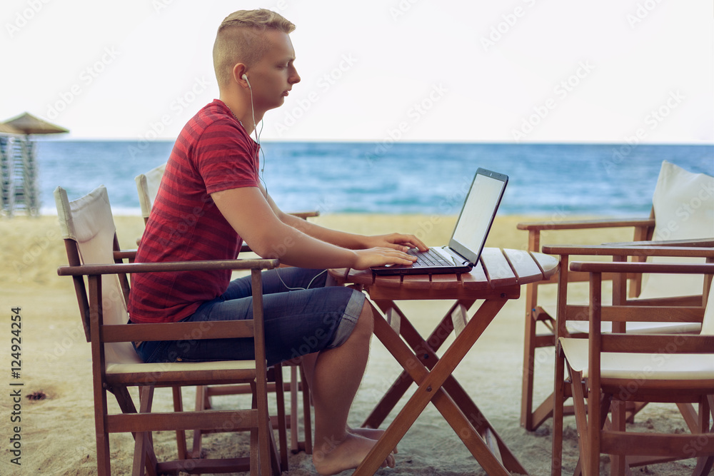 Young businessman.with headphones  on a chair at the table using his laptop on the beach