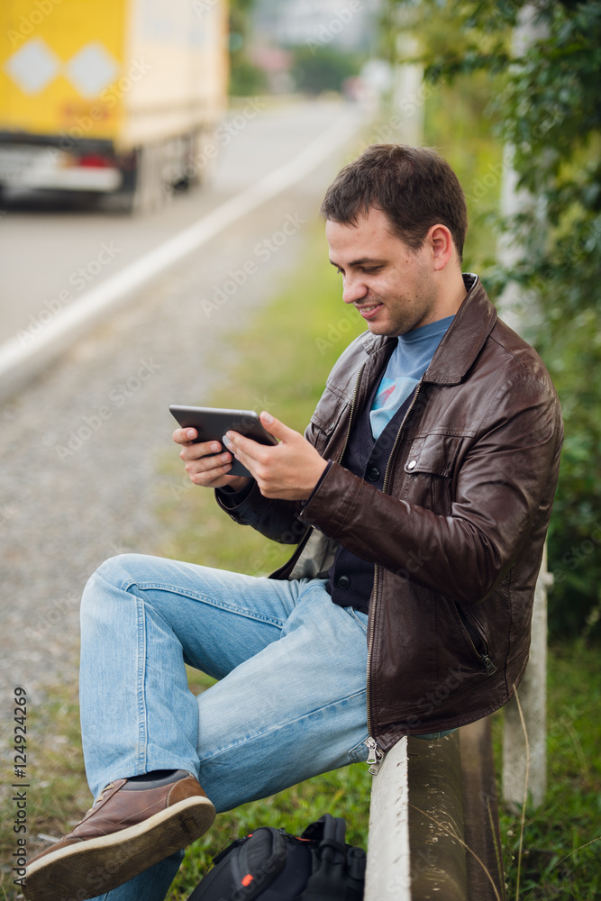 Man orders taxi from his tablet sitting on a road fence Man orders taxi from his tablet sitting on a road fence
