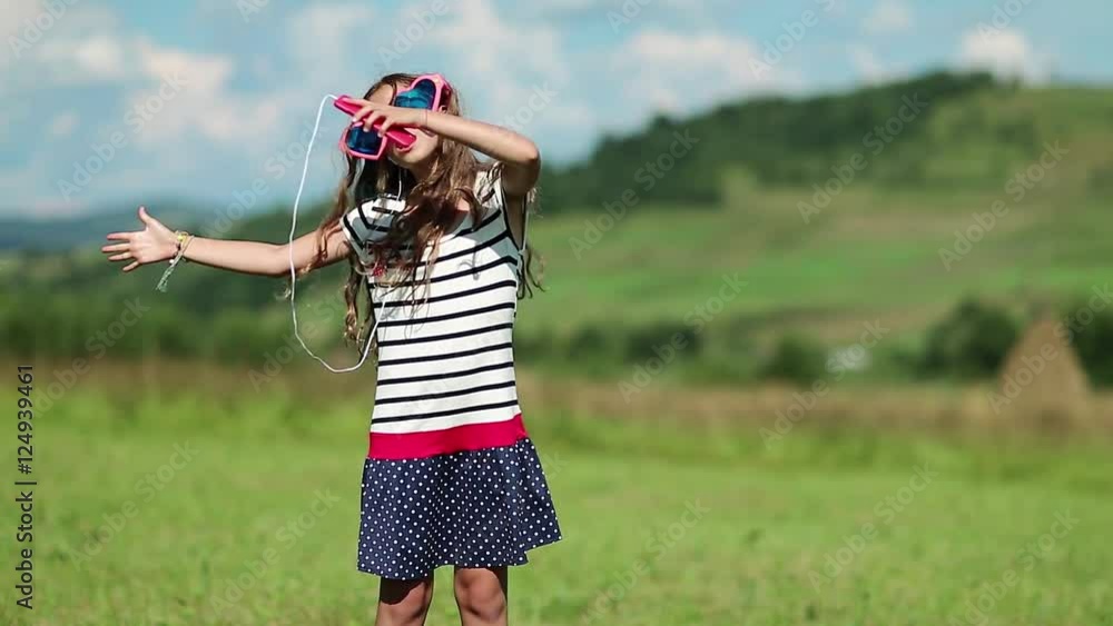 Beautiful girl with smartphone stands on the meadow, listens to music, dances and sings a song. Girl in big glasses with smartphone listens to music and dances on green lawn, and also sings a song Beautiful girl with smartphone stands on the meadow, listens to music, dances and sings a song. Girl in big glasses with smartphone listens to music and dances on green lawn, and also sings a song