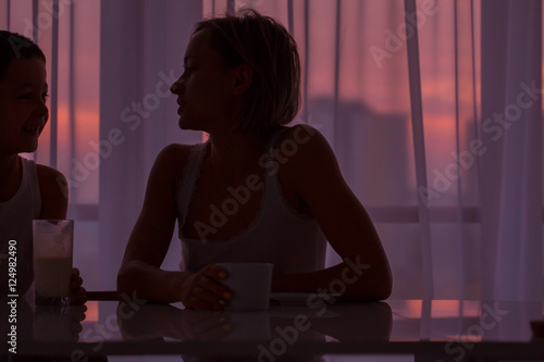 Woman and her son in the morning. Mom and her son are having breakfast, smiling and looking at the sunrise sitting near the window in their home and having a perfect cozy morning.