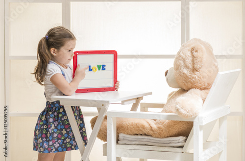Cute toddler playing teacher role game with her toy. Happy little girl holding magnetic board with colored magnetic letters written 
