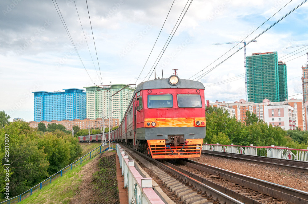 Fototapeta premium Red and grey suburban electric train moves towards on railroad turn vanishing against skyline background. Moscow, Russia. 