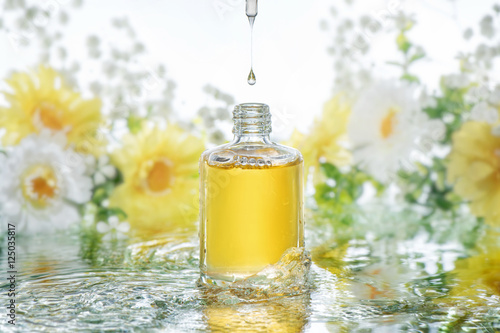 The bottle of moisturizing serum stands in the water waves on the nature blur flowers background. Pipette with drop of serum above the bottle