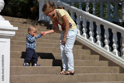 Baby in a plaid shirt making his first steps on a ladder, mother holding his hands supporting by learning to walk