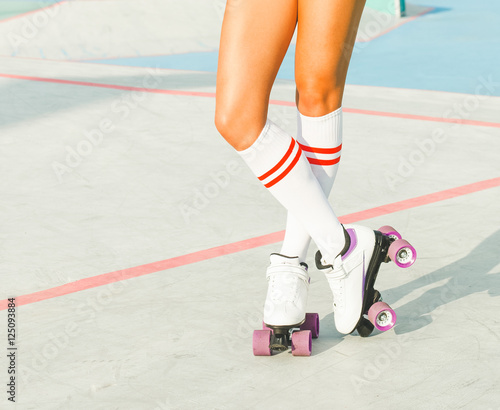 Beautiful long-legged girl posing on a vintage roller skates in denim shorts and white T-shirt in the skate park on a warm summer evening. Part of body.