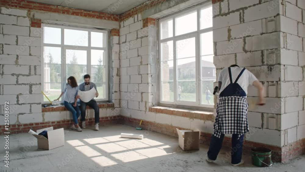 Worker plastering wall with paint roller in empty room of new house while couple sitting and discussing interior plan with digital tablet and paper model Worker plastering wall with paint roller in empty room of new house while couple sitting and discussing interior plan with digital tablet and paper model