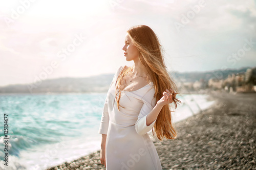 Outdoor summer portrait of young pretty tourist woman with great hair looking to the ocean at europe beach, enjoy her freedom and fresh air, wearing stylish white dress.