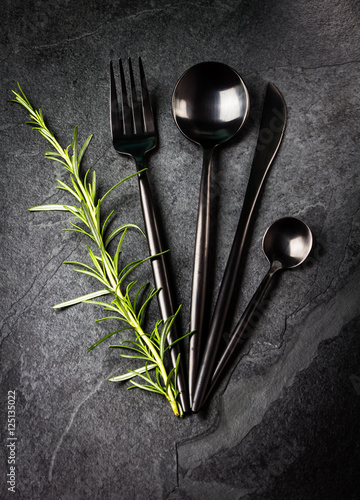 Set of cutlery and rosemary on black stone slate background.