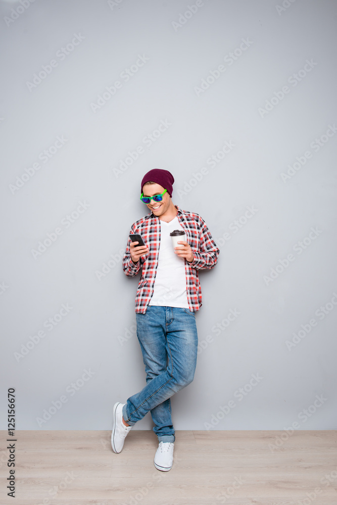 Fototapeta premium Young man in cap and glasses holding cup of coffee and typing sm