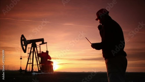 Oil Drilling field ,pumpjack silhouette with setting sun and worker using laptop . Lens Flare.