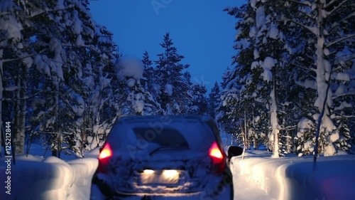 Night winter forest. A lot of snow on the ground and the branches of trees. Snowfall. A car (unrecognizable brand and registration number) with headlights on slowly driving on a forest road