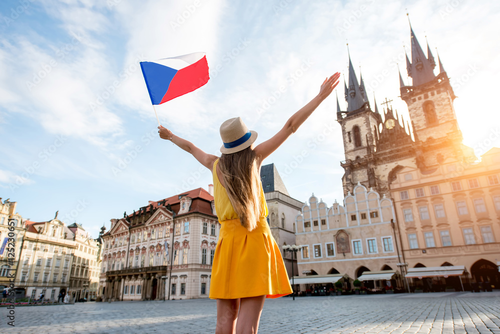 Obraz premium Młoda turystka kobieta ubrana na żółto, trzymając czeską flagę na rynku starego miasta w Pradze. Wspaniałe wakacje w Czechach