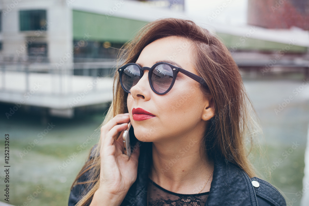 Close-up portrait of a beautiful Caucasian woman with red lipstick. Female is having mobile phone conversation, discussing work tasks. Young female in hipster sunglasses speaks by phone Close-up portrait of a beautiful Caucasian woman with red lipstick. Female is having mobile phone conversation, discussing work tasks. Young female in hipster sunglasses speaks by phone