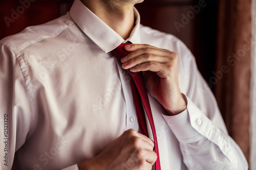 People, business, fashion and clothing concept - close up of man in shirt dressing up and adjusting tie on neck at home. Businessman putting on a tie. Man putting on necktie. 