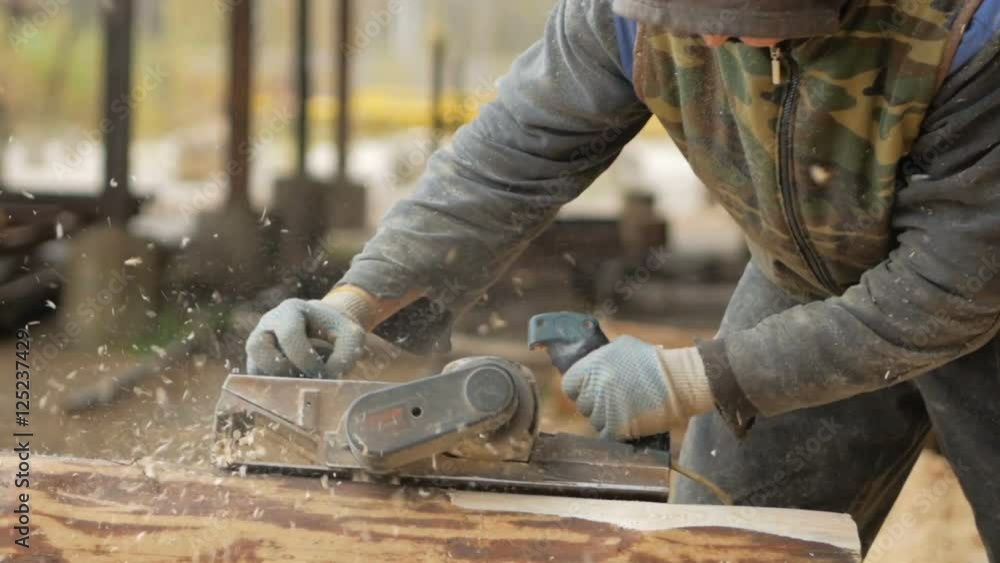 Construction worker planing a piece of wood for a building project. Construction grinds electric hand plane. Against the background of the factory premises, slow motion Construction worker planing a piece of wood for a building project. Construction grinds electric hand plane. Against the background of the factory premises, slow motion