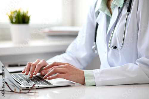 Close up of  unknown female doctor sitting  at the table near the window in hospital and typing at laptop computer