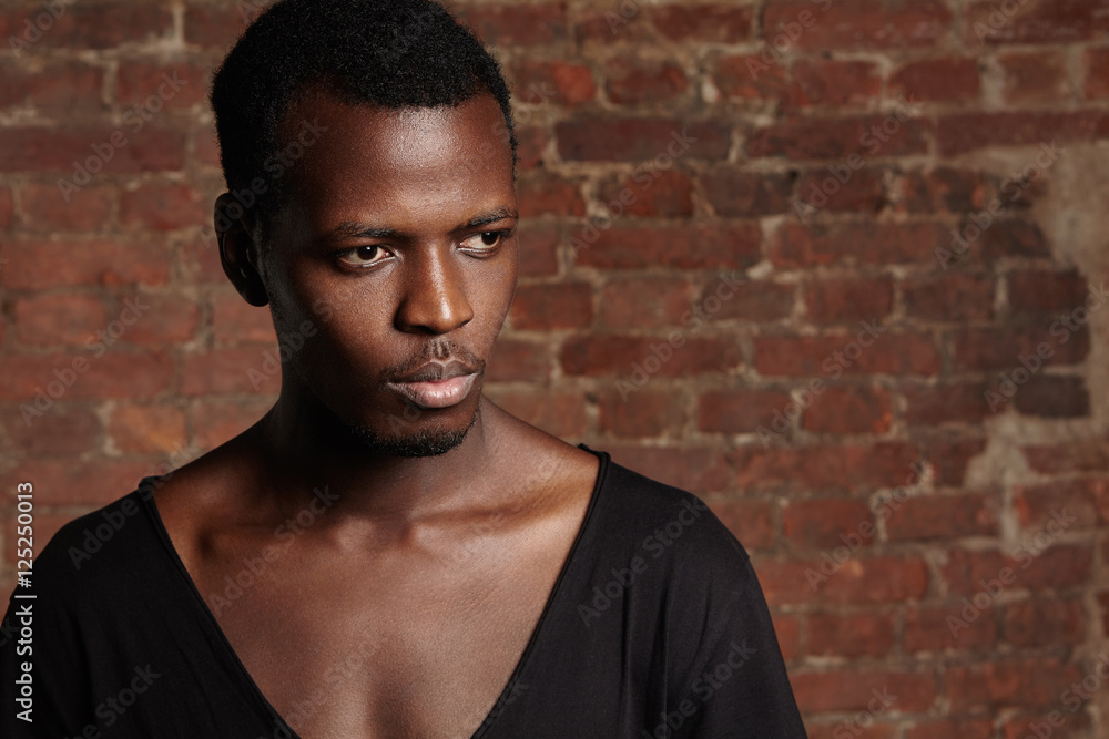 Indoor shot of young African male model with athletic body wearing black stylish T-shirt with scooped neck standing against red-brick wall with copy space for your text or promotional content Indoor shot of young African male model with athletic body wearing black stylish T-shirt with scooped neck standing against red-brick wall with copy space for your text or promotional content