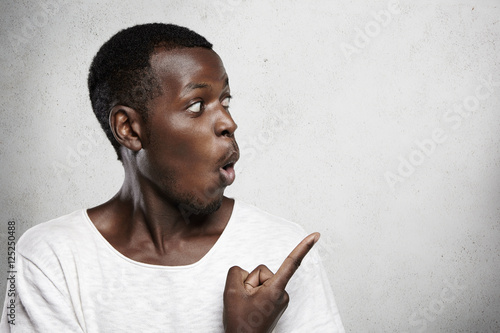 Advertising concept. Midsection of young dark-skinned customer dressed casually, pointing his index finger at blank copy space wall, looking surprised with big sale, opening his mouth widely