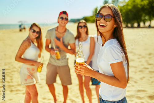 Smiling young girl with beer. other while people barbecuing in the background. People enjoy summer time. Fun at the beach. Great summer mood. sunny day       © maxbelchenko