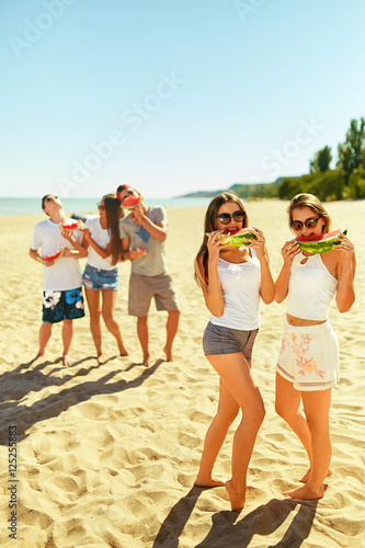 Two young girls eating watermelon on the beach. The other guys in the background. People enjoy summer time. Fun at the beach. Great summer mood. sunny day  