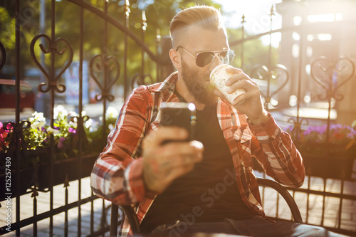 Handsome young man holding mobile phone and drinking coffee while sitting in the rest area of the office. Man dressed in a trendy hipster clothing. 