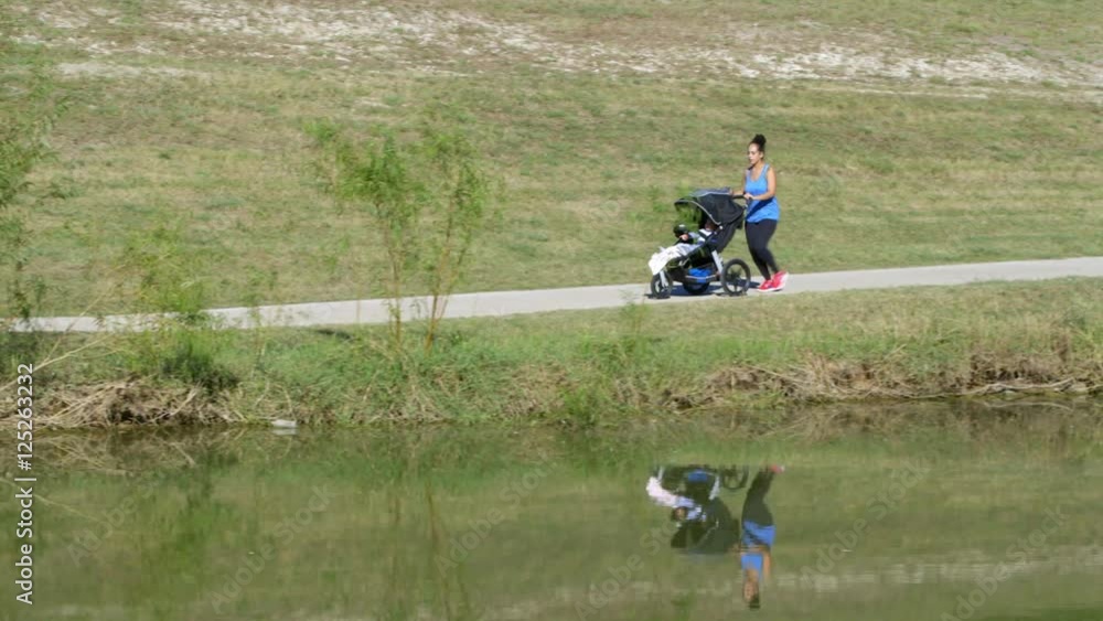 Making time for exercise this busy mom jogs along a lakeside path and across a small bridge pushing her toddler in a three wheel jogging stroller. Making time for exercise this busy mom jogs along a lakeside path and across a small bridge pushing her toddler in a three wheel jogging stroller.