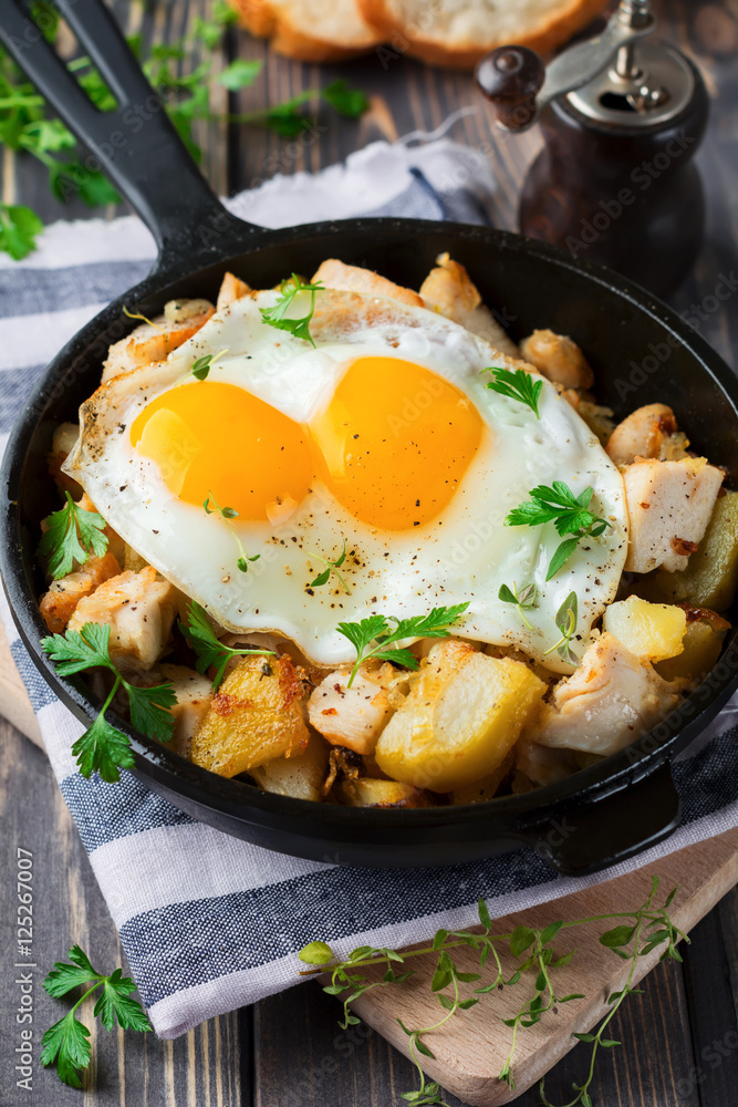 Hearty breakfast. Hash brown potatoes, chicken, onion, parsley, oregano and a fried egg in a frying pan on the old wooden background. Selective focus. Hearty breakfast. Hash brown potatoes, chicken, onion, parsley, oregano and a fried egg in a frying pan on the old wooden background. Selective focus.