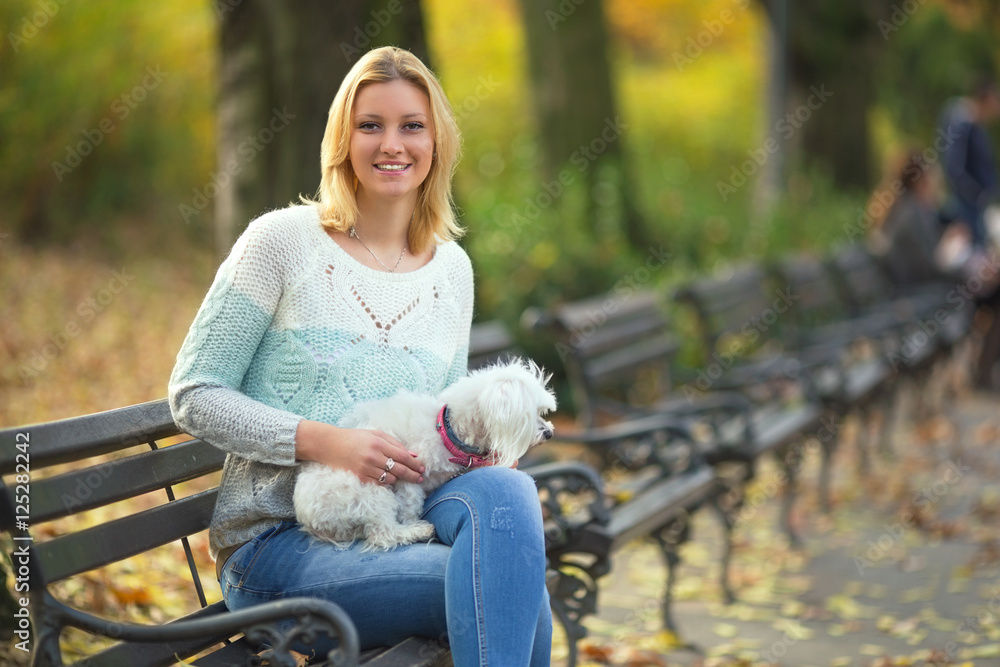 Young woman is sitting on a bench while walking her dog in a park. She is smiling and looking at camera. Young woman is sitting on a bench while walking her dog in a park. She is smiling and looking at camera.
