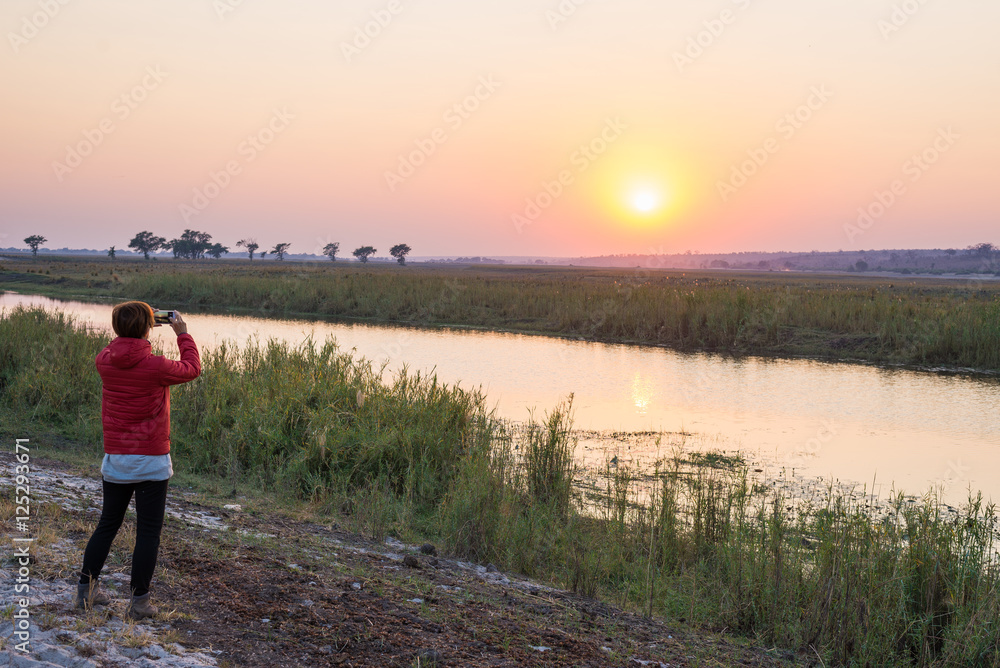 Tourist taking photo with smartphone at majestic sunset over Chobe River, Namibia Botswana border, Africa. Natural colors, rear view.