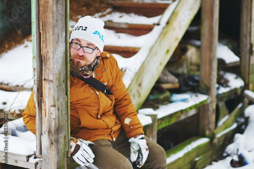 Young bearded man in glasses sitting on the steps of the old wooden stairs in the winter, closeup.