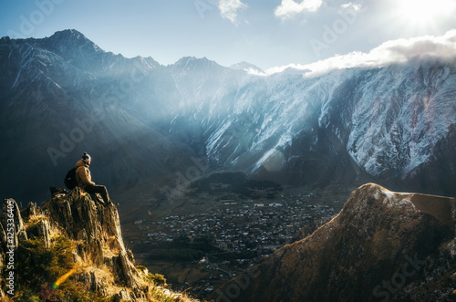 Young tourist in bright hat, black trousers with a backpack sit on cliff's edge and looking at the misty mountain village and glacier at sunrise, Stepantsminda, Georgia