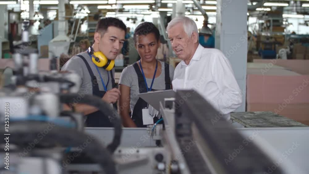 Elderly factory supervisor talking with female Latin-American worker and her Asian colleague about new machinery Elderly factory supervisor talking with female Latin-American worker and her Asian colleague about new machinery