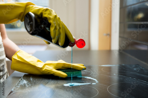 Housewife in rubber protective cleaning and polish electric cooker. Hands pouring cleaning agent on Black surface of kitchen top