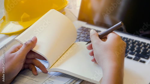 hand of architect holding pen writing on daily ,with laptop computer,  and blueprint, and helmet,soft focus background