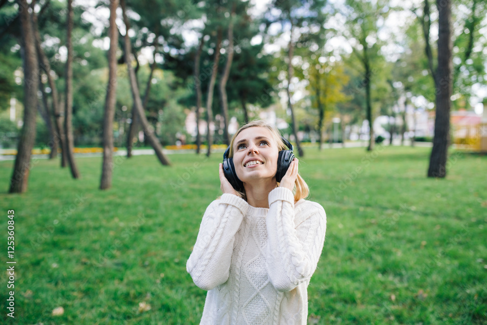 Young beautiful woman enjoying the music outdoor. Happy woman in white sweater with headphones listening to music in the park Young beautiful woman enjoying the music outdoor. Happy woman in white sweater with headphones listening to music in the park