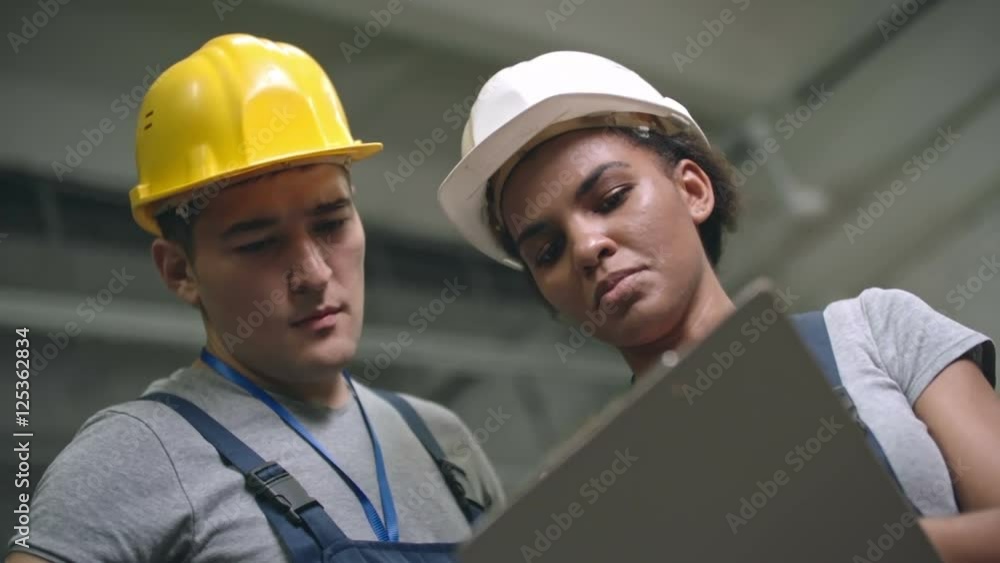 Low angle shot of female Latin-American factory worker with safety helmet holding clipboard and discussing work with Asian male colleague Low angle shot of female Latin-American factory worker with safety helmet holding clipboard and discussing work with Asian male colleague