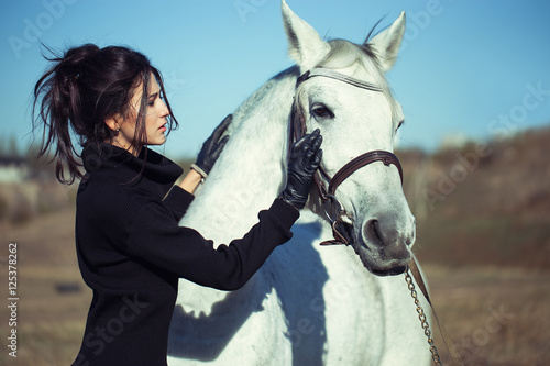  young beautiful girl in the field with white horse. Fashion portrait of brunette model, italian appearance, in casual black pullover. perfect make up, hairdo and stylish look 